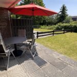 Halb überdachte Terrasse mit rundem Gartentisch und vier Stühlen – zusätzlicher Sonnenschirm für Schatten im sonnigen Garten mit Südost-Ausrichtung.