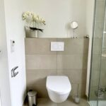 Die Toilette des Badezimmers ist wandhängend montiert. Auf dem Mauervorsprung hinter der Toilette steht eine Blume und ein Kosmetikspiegel.